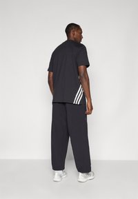 T-shirt noir à manches courtes avec des rayures blanches sur les côtés, associé à un pantalon noir ample et des baskets blanches.