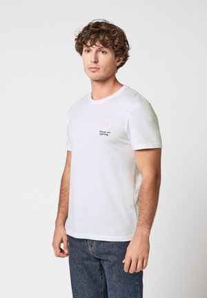 Joven con cabello rizado que lleva una camiseta blanca de manga corta con un pequeño diseño floral de texto, combinada con jeans de color azul oscuro, sobre un fondo liso.