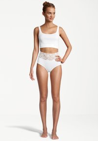 Frau in weißem, cropped Tanktop und hochgeschnittenen weißen Unterhosen mit Spitzenakzenten, barfuß vor einem schlichten weißen Hintergrund stehend.