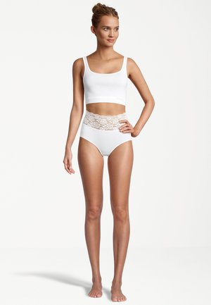 Frau in weißem, cropped Tanktop und hochgeschnittenen weißen Unterhosen mit Spitzenakzenten, barfuß vor einem schlichten weißen Hintergrund stehend.