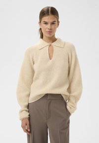 Krēmkrāsas adīta džemperis ar ribotu tekstūru, ar apkakles izgriezumu un atslēgas caurumu priekšpusē, saskaņots ar brūniem biksēm.