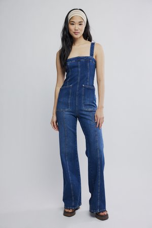 Mlada ženska, ki nosi modri jeans kombinezon s širokimi hlačnicami in kvadratnim izrezom, kombiniran s rjavimi sandali na platformo in bež trakcem za lase.