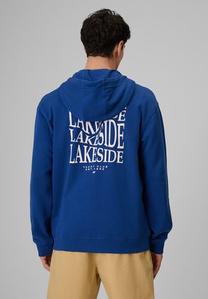 Mężczyzna w niebieskiej bluzie z kapturem z napisem „LAKESIDE Yacht Club EST. 1985” na plecach, w połączeniu z beżowymi szortami.