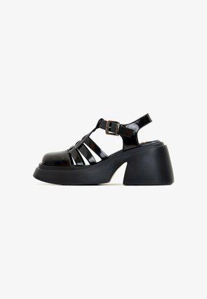 Schwarze lackierte Ledersandalen mit geschlossenem Zehenbereich, ausgeschnittenen Riemen, verstellbarem Schnallenverschluss und einem massiven schwarzen Plateauschuh.
