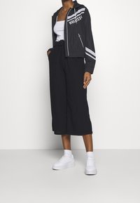 Černá zkrácená bunda s bílými pruhy a textem "Hollister", doplněná volnými černými kalhotami a bílými teniskami. Hladký materiál, volný střih.