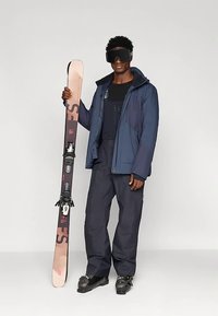 Skiausrüstung: eine Person in einer dunkelblauen Jacke, schwarzen Hosen, hält hellbraune Skier mit schwarzen Bindungen, trägt eine große schwarze Skibrille.