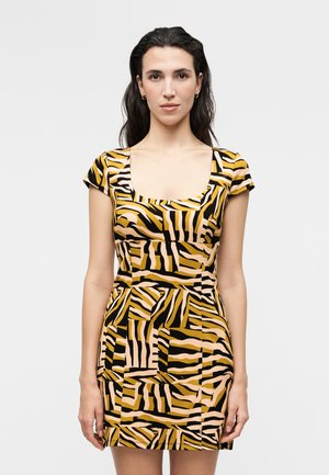 Žena nosi šaty s krátkym rukávom a štvorcovým výstrihom s odvážnymi čiernymi, žltými a béžovými abstraktnými zebrovitými pruhmi na svetlom pozadí.