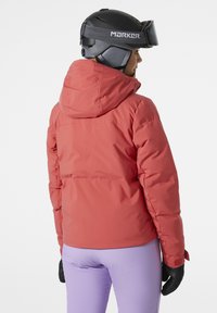 Rote isolierte Skijacke mit Kapuze, strukturierter Stoff und taillierter Passform, kombiniert mit lila Skihosen und schwarzen Handschuhen.
