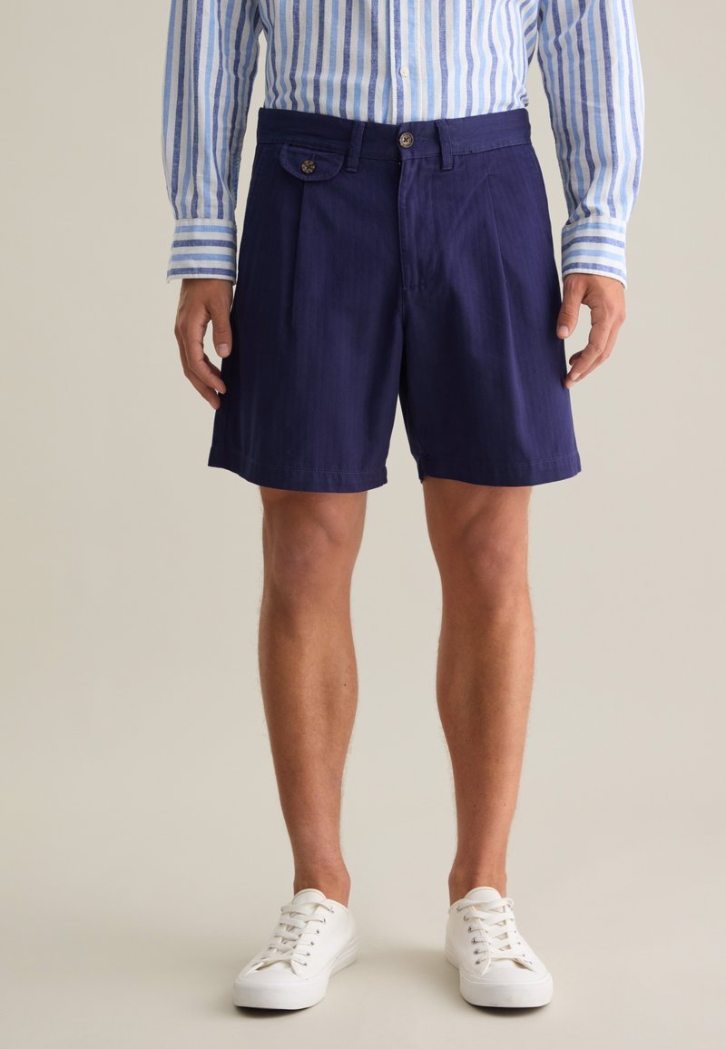 Marineblaue Shorts aus leichtem Material, mit flacher Vorderseite, einem Knopf und seitlichen Taschen, die bis zum Knie reichen.