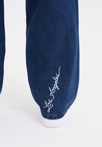 Blå denimbyxor med vit broderad text "Los Angeles" vid fållen, med en avslappnad passform och elastiska nederkanter vid anklarna.