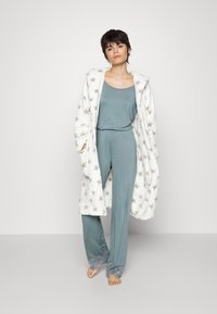 Robe en polaire blanche avec des étoiles dorées, portée par-dessus un ensemble de vêtements d'intérieur bleu sarcelle. Le pantalon a des ourlets à motif floral, avec une coupe décontractée.