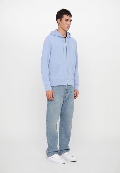 Fato de treino azul claro com fecho éclair, bolso frontal, capuz com cordão e um pequeno logótipo. Usado com jeans azul claro e sapatilhas brancas.
