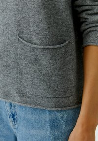 Pull en tricot gris avec une poche avant, un col rond et des poignets côtelés. Le tissu a une texture douce, associé à un jean déchiré bleu clair.