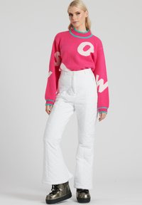 Maglione rosa brillante con stampa "S O W", abbinato a pantaloni bianchi trapuntati e stivaletti metallici. Il maglione presenta un colletto e dei polsini turchesi.