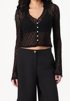 Kvinde iført sort gennemsigtig blonder crop top med lange ærmer og knapper, over en sort bh og højtaljede sorte bukser med knaplukning.