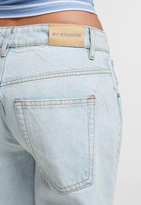 Lichtblauwe denim jeans hebben een klein leren label op de tailleband met "2Y STUDIOS" in reliëf. Zichtbare achterzakken en naaddetails.