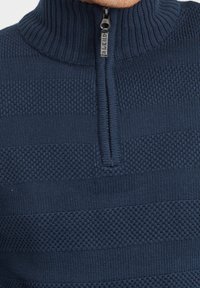 Maglione lavorato a maglia blu navy con colletto alto, caratterizzato da un design con zip a un quarto e strisce orizzontali testurizzate. Accentuato da un tirazip con logo.