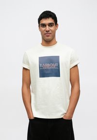 Kremowy t-shirt z krótkim rękawem z kwadratowym nadrukiem z napisem "BARBOUR INTERNATIONAL" na czerwono na niebieskim tle.