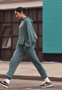 Turkis hoodie og matchende joggere med en løs pasform og lommer. Grønne sneakers med lyserøde accenter, parret med hvide sokker på en bygade.
