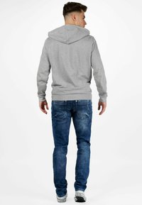 Haut à capuche gris clair avec une capuche, doté de manches longues et de poignets côtelés, accompagné de jeans en denim bleu. Des baskets réfléchissantes complètent la tenue.