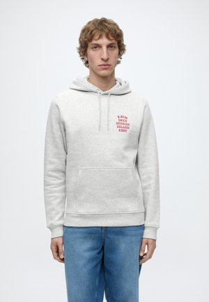 Jeune homme aux cheveux bouclés portant un sweat à capuche gris clair avec un texte rouge et un jean bleu, debout sur fond blanc.