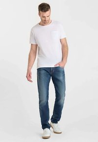 T-shirt blanc à col rond avec petite poche poitrine, associé à un jean en denim bleu moyen et des baskets blanches. Coupe décontractée, design minimaliste.