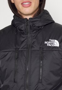Juodas gobtuvas striukė su išklotuoju dizainu, turinti dvi užtrauktukų kišenes ir balta "The North Face" logotipą viršutiniame kairiajame šone.