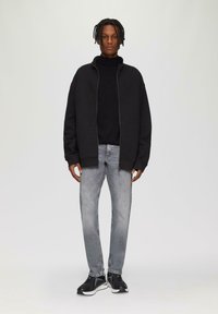 Veste zippée noire sur un col roulé noir, associée à un jean gris clair et des baskets noires. Tissu lisse, coupe décontractée, design minimaliste.