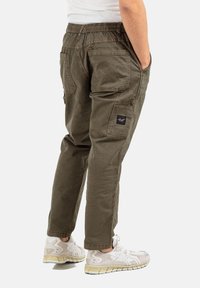 Man wearing olive green cargo pants with back and side pockets and white sneakers, standing with hands in pockets.