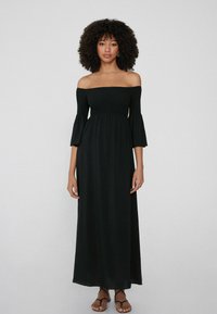 Vestido maxi negro sin mangas con mangas acampanadas, cuerpo fruncido, falda fluida y tejido ligero, combinado con sandalias negras.