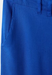 Gros plan sur un pantalon en tissu bleu montrant la poche avant, la ceinture et les passants de ceinture avec des détails de couture visibles.