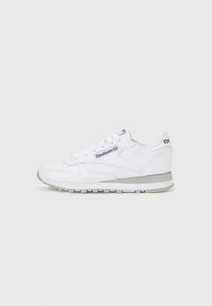 Białe skórzane sneakersy Reebok Classic z szarą podeszwą i czarnym logo Reebok, widoczne z boku na jednolitym jasnoszarym tle.