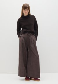 Brauner Strickpullover mit hohem Ausschnitt, kombiniert mit weiten, braunen Karohosen. Das Outfit wird durch burgunderfarbene Sneakers ergänzt.