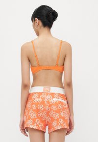 Oranje geribde bralette met dunne bandjes, gecombineerd met oranje shorts met witte bloemenpatronen en een contrasterende witte tailleband.
