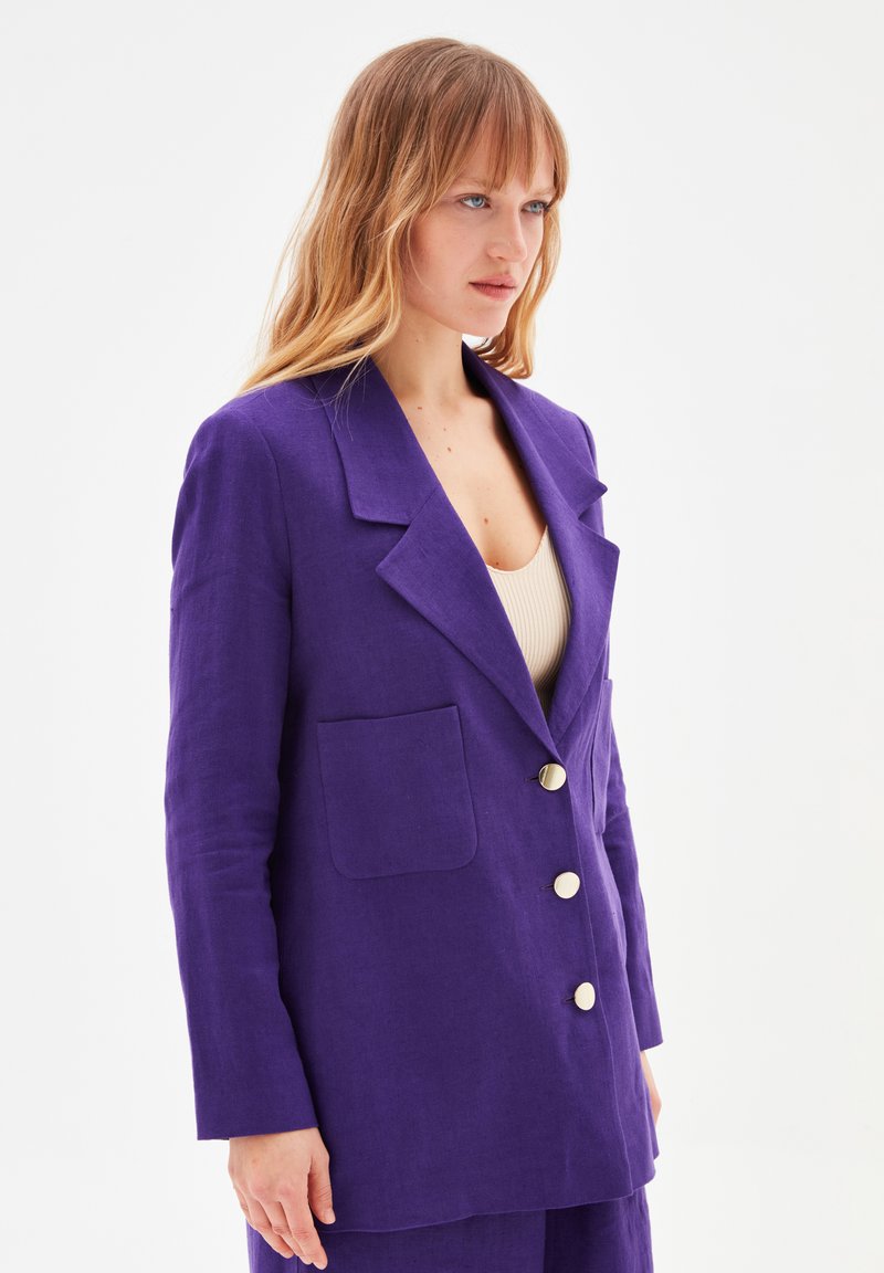Blazer in lino viola con una tasca singola, colletto a rever e tre bottoni dorati. Presenta una vestibilità rilassata con maniche lunghe e una texture morbida.