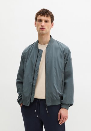 Young man wearing a blue bomber jacket over a beige shirt and navy drawstring pants, standing against a plain white background.