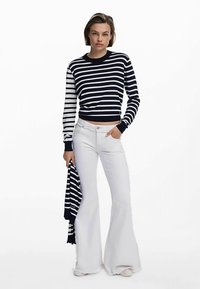 Laivastonsininen ja valkoraidallinen neulepaita, jossa on resorikaulus, yhdistettynä valkoisiin liehukehousuihin. Malli pitää kädessään vastaavan huivin ja seisoo suorassa.