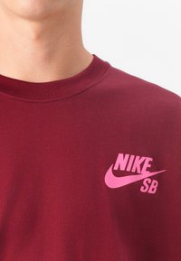 T-shirt en coton marron avec un logo Nike rose bien en vue et le texte SB sur la poitrine gauche. Modèle à col rond avec une texture lisse.