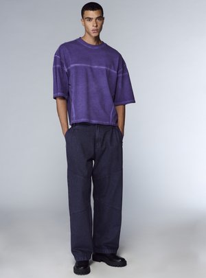 Maglietta oversize viola in cotone con cuciture a contrasto, abbinata a pantaloni di denim scuro con più pannelli e tasche, completata da stivali neri.
