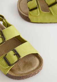 Gröna mockasandaler med två justerbara remmar, bronsfärgad spännen och en texturerad tan inläggssula på en brun, greppvänlig sula.