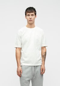 Λευκό βαμβακερό t-shirt με κοντά μανίκια και στρογγυλή λαιμόκοψη, που διαθέτει μια διακριτική λεπτομέρεια λογοτύπου στο στήθος. Το μοντέλο φορά γκρίζες φόρμες.