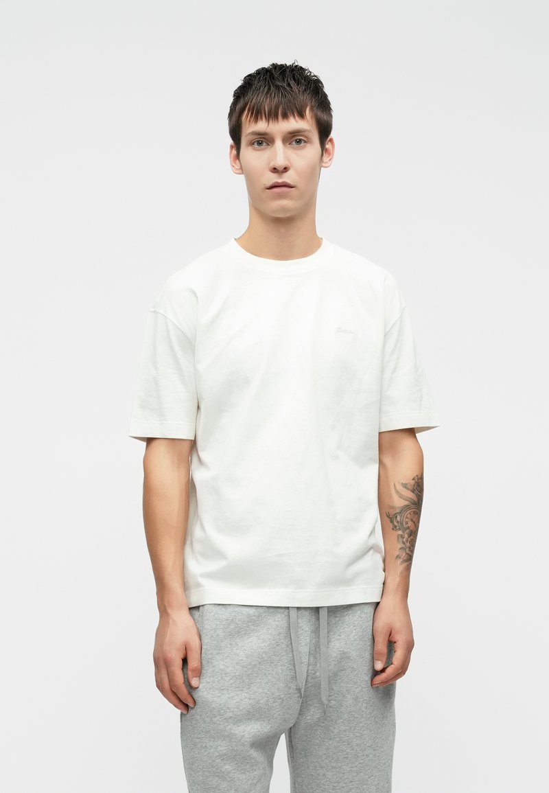 Λευκό βαμβακερό t-shirt με κοντά μανίκια και στρογγυλή λαιμόκοψη, που διαθέτει μια διακριτική λεπτομέρεια λογοτύπου στο στήθος. Το μοντέλο φορά γκρίζες φόρμες.