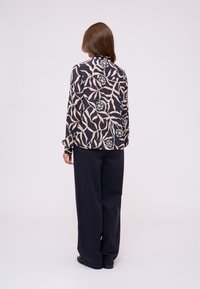 Długa bluzka z długim rękawem w kolorze granatowym z beżowym abstrakcyjnym wzorem liści i okrągłymi dodatkami; zestawiona z szerokimi, ciemnymi spodniami, widok od tyłu.