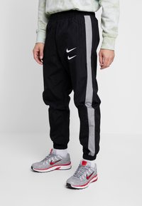 Jogging en nylon noir avec des rayures latérales grises et une taille élastique. Comprend le logo Nike en blanc sur la cuisse gauche. Associé à des baskets grises.
