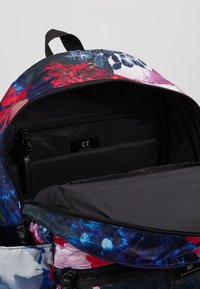 Reppu, jossa on eloisa kukkakuosi, avoinna mustan sisäosan ja vetoketjullisen taskun näyttämiseksi.