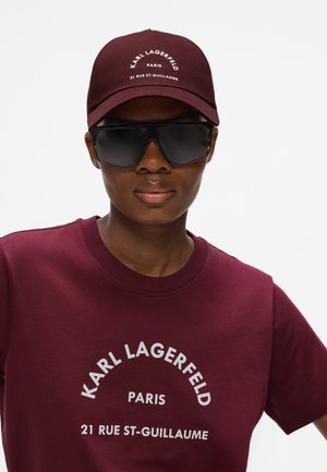 Person wearing dark oversized sunglasses, burgundy cap, and matching t-shirt with "Karl Lagerfeld Paris 21 Rue St-Guillaume" text.