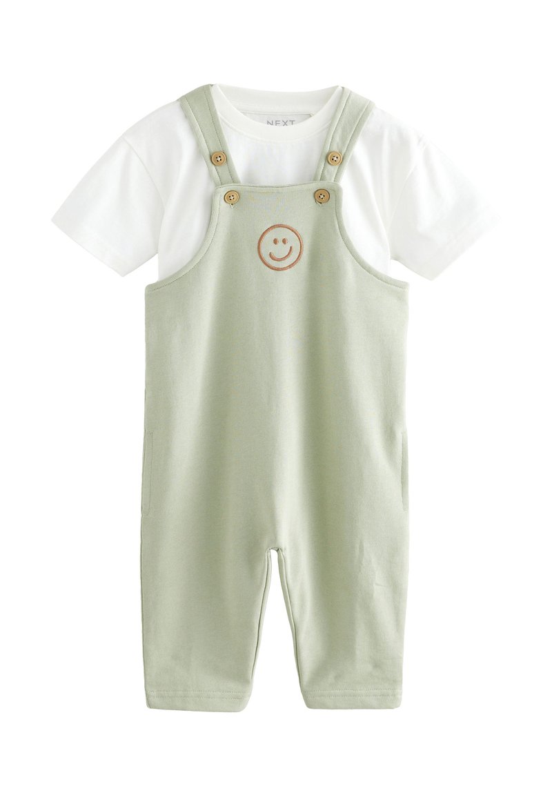 Gaiši zaļas kopbikses ar regulējamām lencēm, koka pogām un smaidošu sejas emblēmu priekšpusē, kopā ar baltu īsroku kreklu.