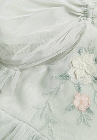 Ljust grönt tyg med genomskinlig textur, prytt med blommönster i vitt och ljusrosa, med rynkade detaljer längs kanterna.