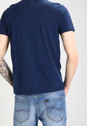 Homme portant un t-shirt bleu foncé et un jean bleu clair, avec une manchette de tatouage sur son bras gauche, tourné dos à la caméra.