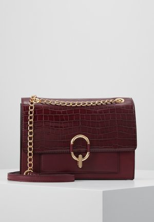 Sac à main en cuir bordeaux avec texture crocodile, bandoulière chaîne dorée et fermoir ovale doré, présenté sur un piédestal blanc.
